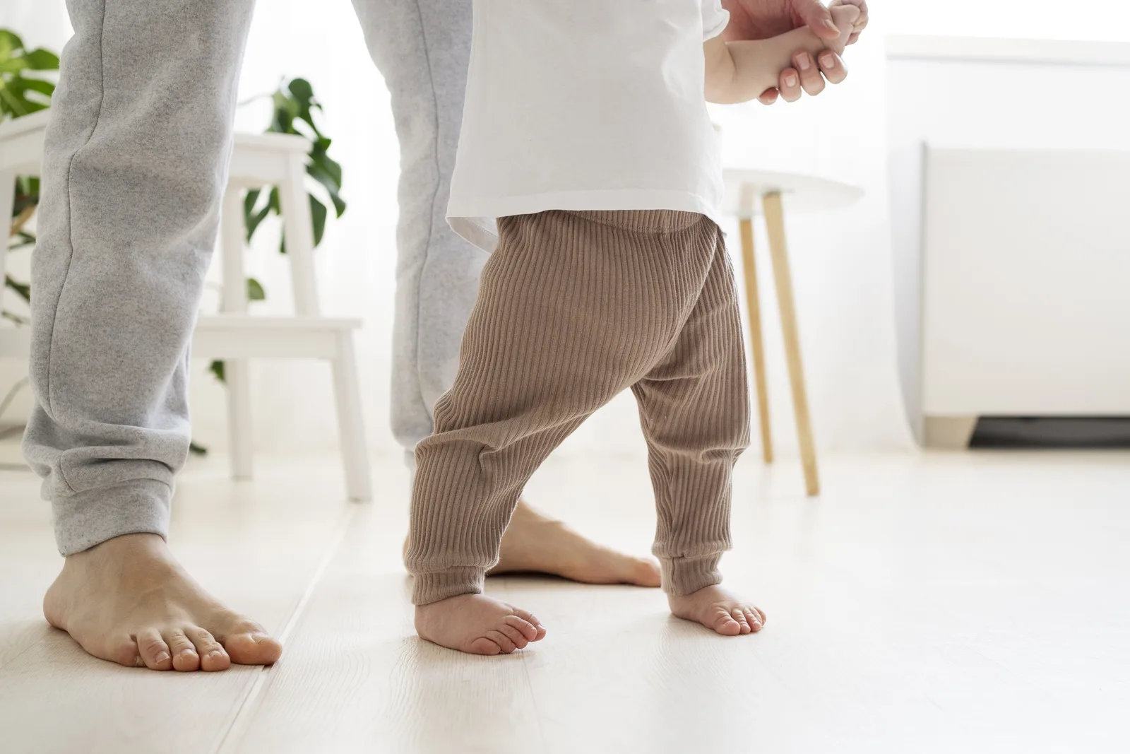 Los pies descalzos de un bebé que empieza a andar sobre un suelo claro, junto a los pies de un adulto que lo lleva de la mano