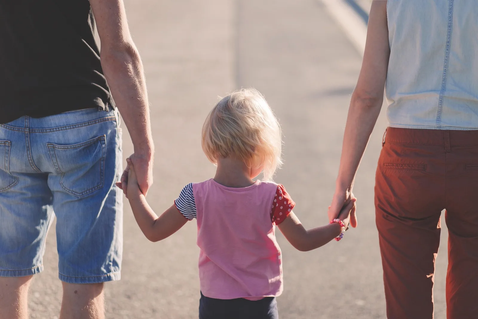 Una niña pequeña de espaldas, caminando de la mano de su padre y su madre